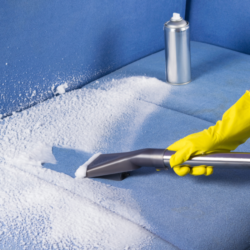 A rubber gloved hand holding a vacuum cleaner and cleaning foam cleaner off a blue sofa