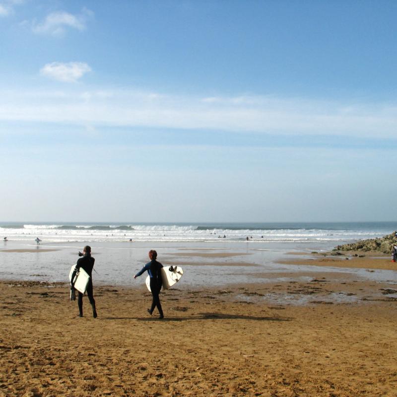 Two people holding surfboards walking away from us heading towards the surf