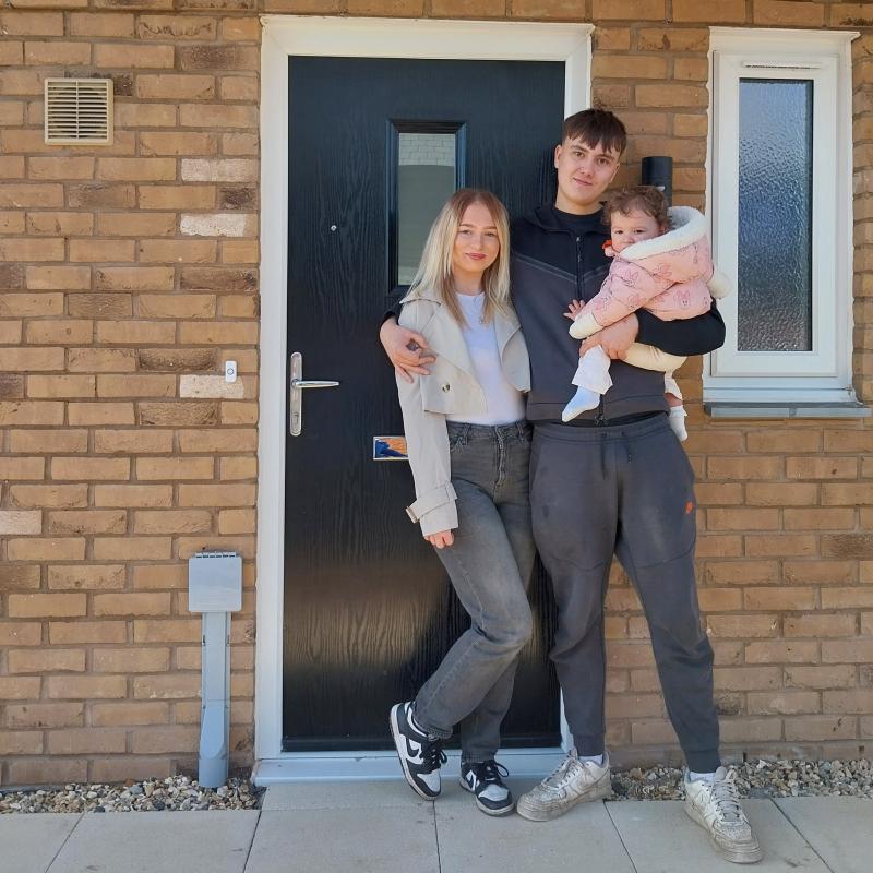 A young man holding a female child with his arms around a young woman stood outside their front door